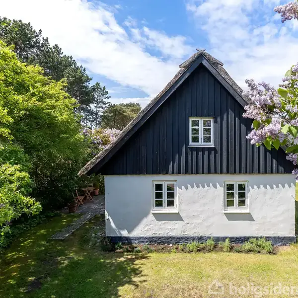 Et sort-hvidt hus med skråtag står i en frodig have, omgivet af træer og blomstrende syrener under en klar blå himmel.