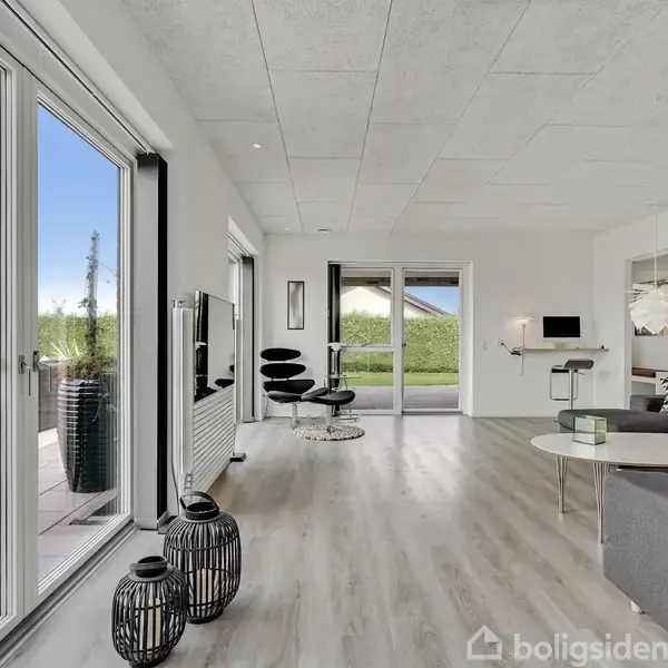 Stue med grå sofa og runde borde, åbner mod terrasse med grønne omgivelser. Store vinduer giver lys, vægmonteret tv ses foran moderne stole. Minimalistisk og lys indretning.