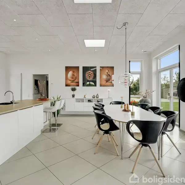 Et moderne køkken-alrum med en lang, hvid spisebord, sorte stole og indbyggede skabe. Store vinduer og glasdøre leder ud til en have. Tre kunstværker hænger på væggen.