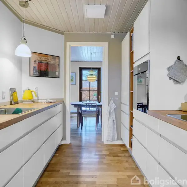 En moderne køkkenindretning med hvide skuffer og træbordplader, hvor en gul lampe og en vask er synlige. Gennem døråbningen ses et spisebord i et tilstødende rum.