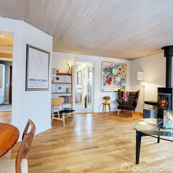 Rundt spisebord i træ står i et lyst rum med trægulv, moderne brændeovn i hjørnet, og kunst på væggene. Åbne døråbninger fører til et køkken og en gang.