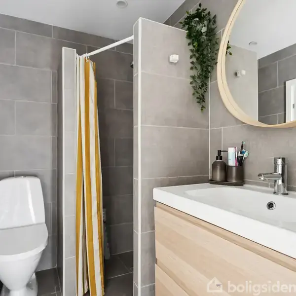 Toilet ved siden af vask med spejl, placeret i moderne badeværelse. Bruseniche med gulv-til-loft-fliser og gulstribet bruseforhæng. Grøn plante hænger over spejlet, skaber et friskt look.