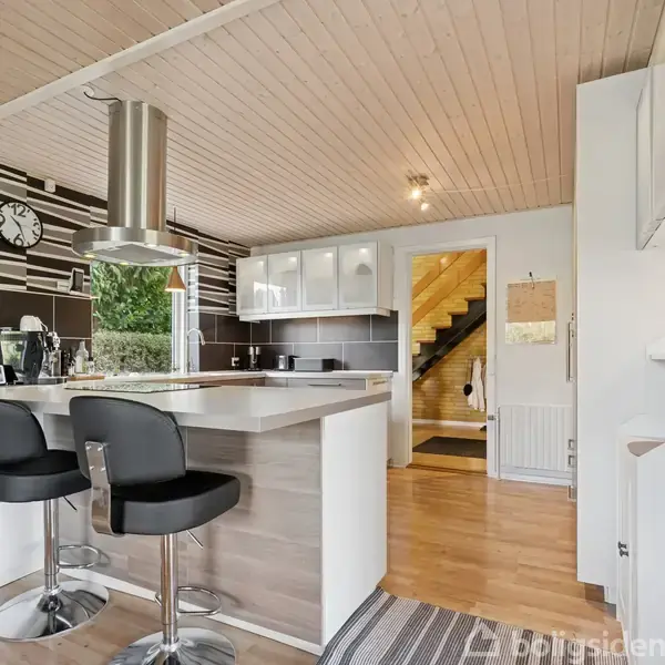 Køkken med træloft har en ø med to sorte stole og emhætte. Skandinavisk stil med stribede vægge, hvidt skab og adgang til gang med trappe. Indbydende rum.