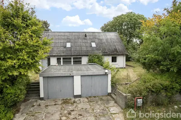 En hvid villa med skråt tag og to garageporte står omgivet af grønne træer og buske. Foran huset er der en flisebelagt indkørsel.