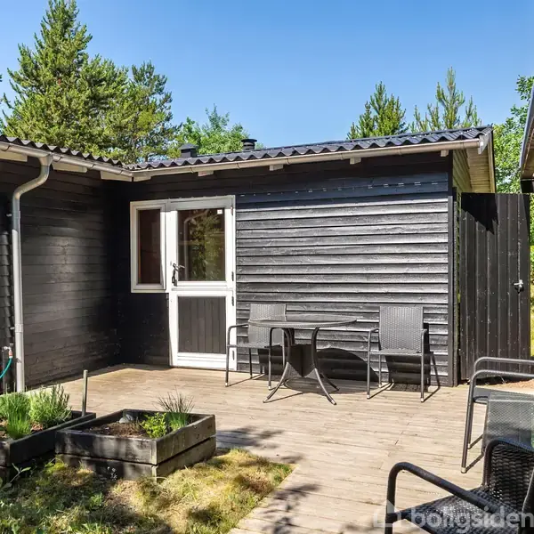 Sortmalet hus med træterrasse. Terrassen har to stole og et bord. Omkring huset er der grønne træer og buske, som skaber en naturlig og afslappende atmosfære.