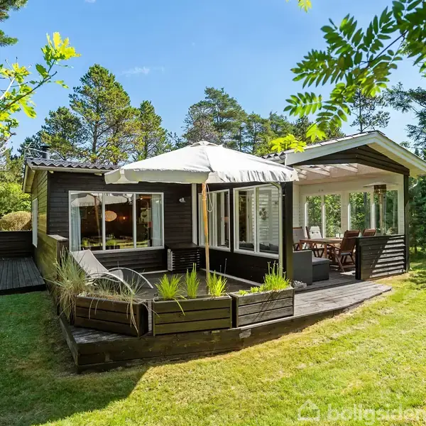 Sommerhus med terrasse og parasol, omgivet af grøn græsplæne og træer under en klar blå himmel.