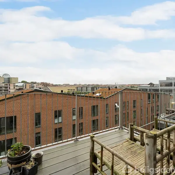 En balkon med planter står stille, omringet af byens røde murstensbygninger og en klar blå himmel. Rails og siddemøbler skaber en hyggelig atmosfære.