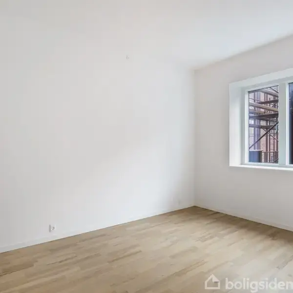 Et tomt rum med trægulv og hvide vægge. Rummet har et stort vindue, der kigger ud mod en rødstensbygning og et stillads, hvilket indikerer opførelse eller renovering udenfor.