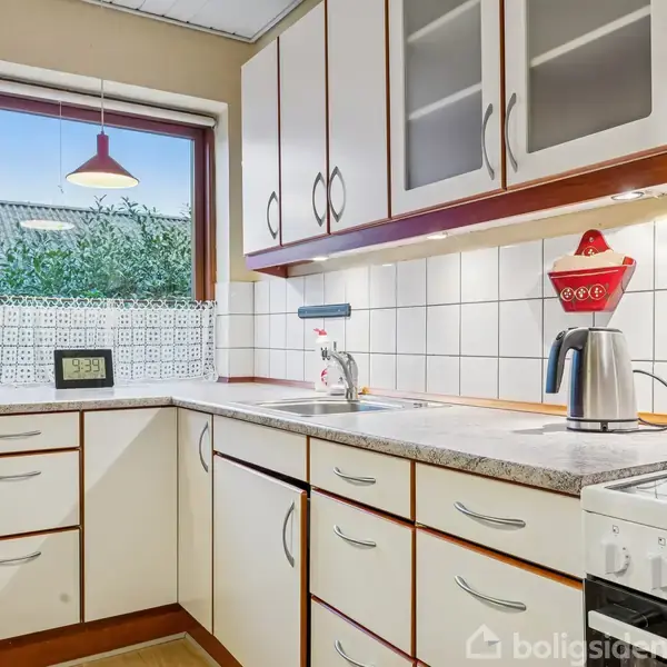 Et hvidt køkken med trækanter har skabe og skuffer. En elkedel står på bordet ved siden af en rød lampeskærm. Et vindue giver udsigt til et hustag og træer.