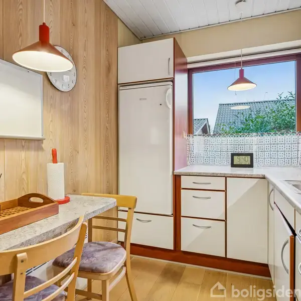 Et køkkenbord står med stole, tæt på et vindue. En bordlampe hænger over bordet. Bag væggen ses et køleskab og skabe. Udenfor ses et hustag og træer.