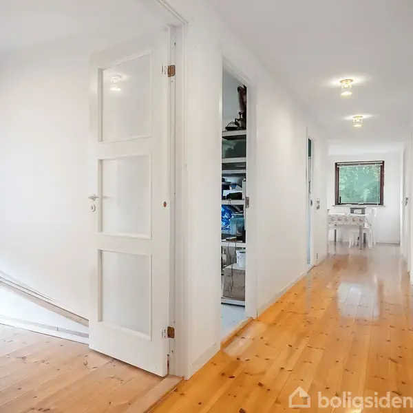 En lang, lys gang med træparketgulv og hvide vægge. Flere døre åbner sig til forskellige rum. I slutningen ses et vindue med grønt udendørs. En stol og et bord står for enden.