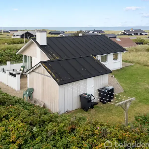 Et hvidt sommerhus med sort tag står roligt i en grøn, kuperet kystnær landskab. Andre sommerhuse kan ses i baggrunden nær havet.