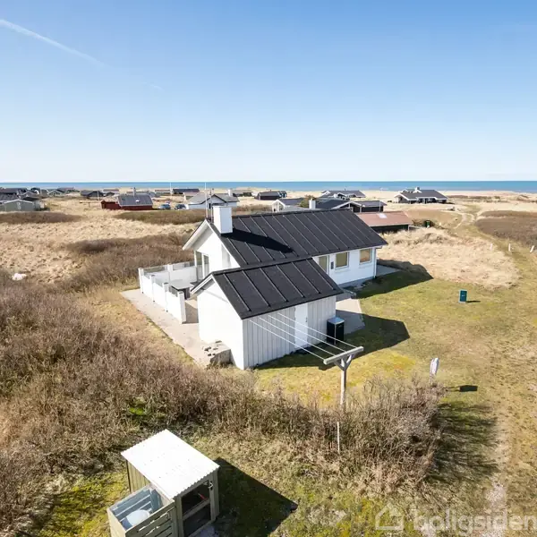 Et hvidt sommerhus med solpaneler ligger blandt sandklitter nær havet. Omkringliggende vegetation består af græs og lave buske. Flere huse ses spredt i det fjerne.