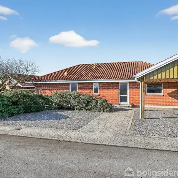 Rødt murstenshus med rødt tegltag står ved siden af en gul træcarport. Huset er omgivet af buske og har en stenbelagt indkørsel, beliggende i et boligkvarter.