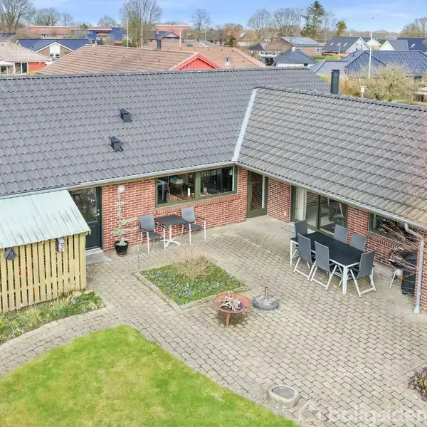 Et murstenshus med mørkt tag har to terrasser med havemøbler. Der er en have med græs og hæk i et villakvarter med andre huse omkring.