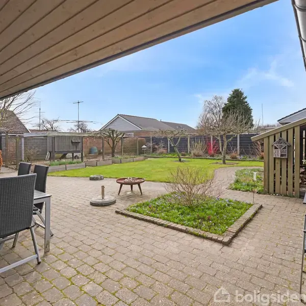 Terrasse med havemøbler står på en flisebelagt overflade. En lille have med græs og buske omkranser området. Hegn og huse ses i baggrunden under en klar blå himmel.