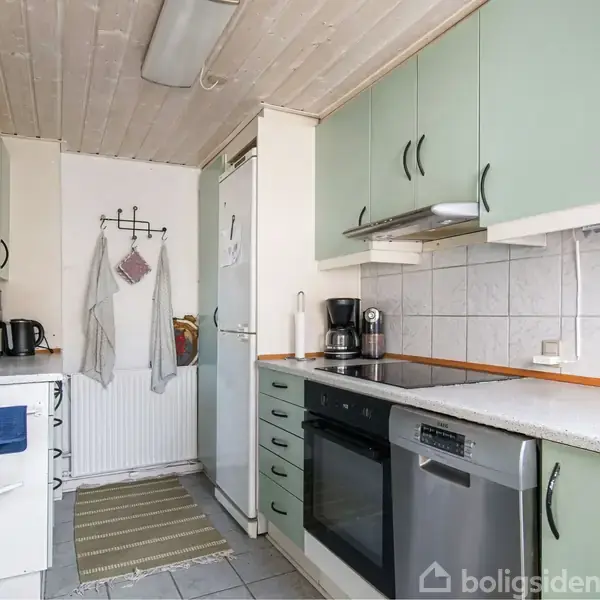 Et køkken med lysegrønne skabe, en opvaskemaskine, ovn, og kaffemaskine. En disk og vask ses på højre side. En tæppebelagt gulvgang fører til et køleskab med håndklæder påført.