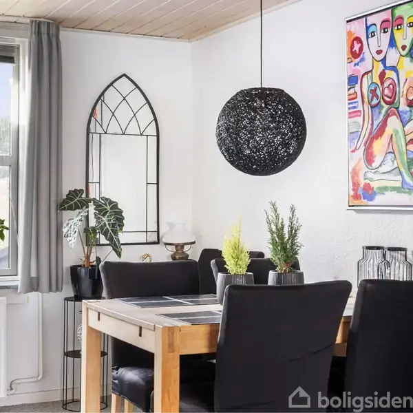 Et spisebord med sorte stole står i et lysfyldt rum. En rund sort lampe hænger over bordet. Der er planter og farverige kunstværker på væggen.