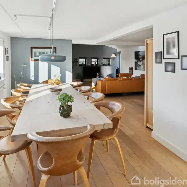 Et langt spisebord med træstole er arrangeret i et lyst spisestueområde. På bordet står planter. I baggrunden ses en brun sofa og et køkkenhjørne med moderne apparater.