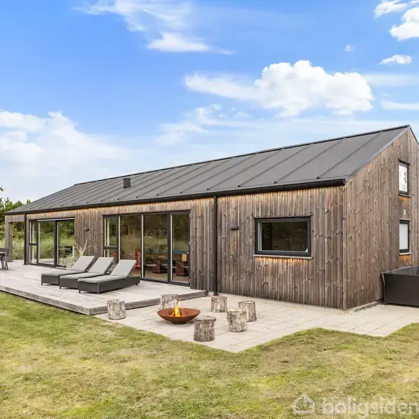 En moderne træbygning med store vinduer står på en flisebelagt terrasse. To liggestole og en ildskål pryder terrassen. Græsplæne og træer omkranser huset under en blå himmel.