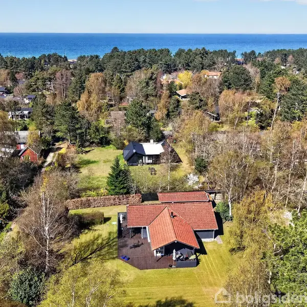 Hus med rødbrun tag, omgivet af stor have og træer, beliggende i skovområde med udsigt til havet i baggrunden. Klar blå himmel over området.