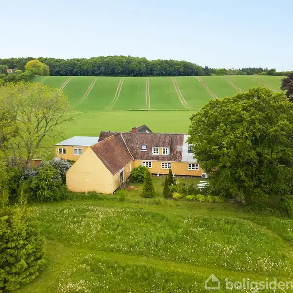 Gult landhus omgivet af frodig have og store træer, beliggende i et grønt, kuperet landskab med marker i baggrunden.
