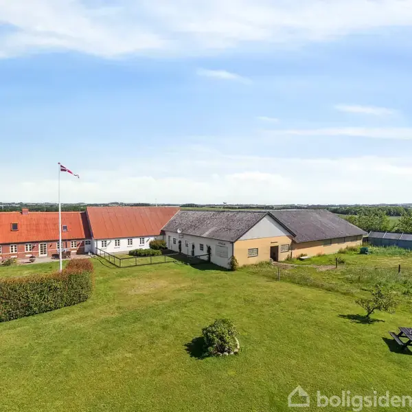 En stor gård med rødt tag og flagstang står stille i et åbent landskab. Gården omgives af græsplæne, hegn og spredte træer under en klar blå himmel.