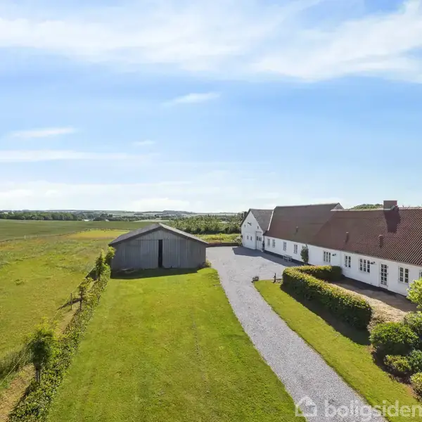 Landhus står stille ved siden af en grussti omkranset af græs og buske; en udbygning ligger ved siden af, omgivet af åbne marker under en blå himmel. En hund ses til venstre.