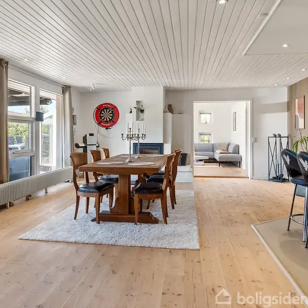 Et spisebord med otte stole står på et lyst tæppe i en moderne spisestue med trægulv og hvide lofter. Store vinduer til venstre giver udsigt til en terrasse.