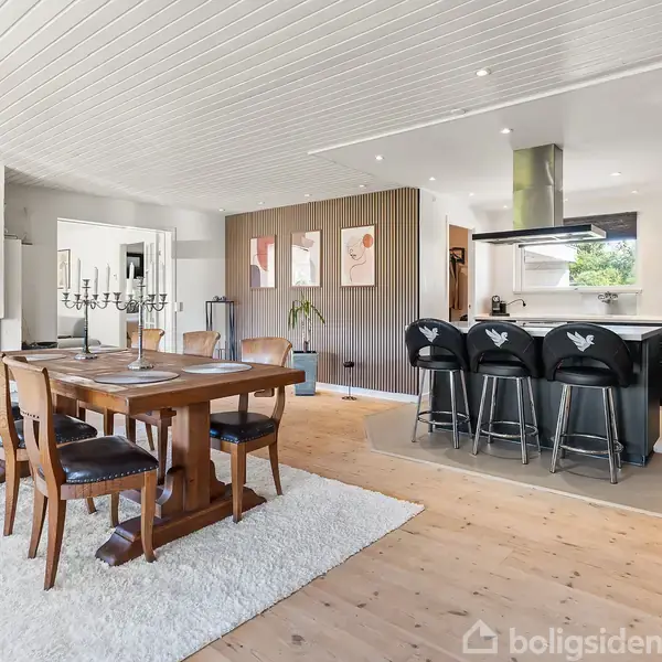 Et spisebord af træ med stole står på et hvidt tæppe. I baggrunden ses et moderne køkken med barstole ved en ø. Rum med lyse trægulve og hvidt loft.