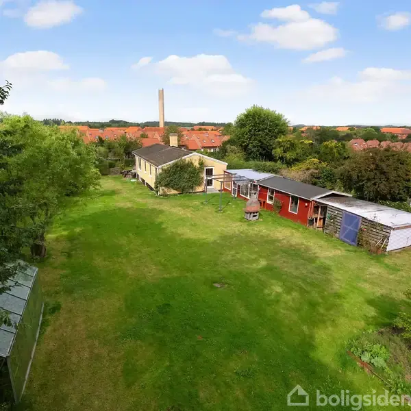 Et langt, rødt skur står på en stor, grøn græsplæne i et åbent, landligt område med træer og huse i baggrunden under en blå himmel med skyer.