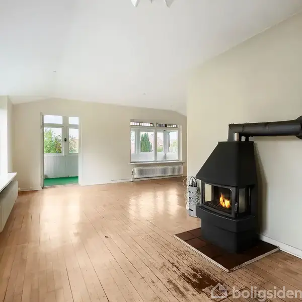 Brændeovn varmer en lys stue med trægulv og store vinduer. Rummet har hvide vægge, en radiator under vinduet, og åbner mod et andet rum med grønne gulve.