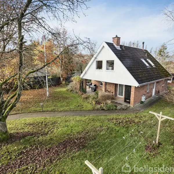 En rød murstenshus med hvid gavl og mørkt tag står på en græsplæne ved siden af en trægyngestativ. Det er omringet af bartræer i en have om efteråret.