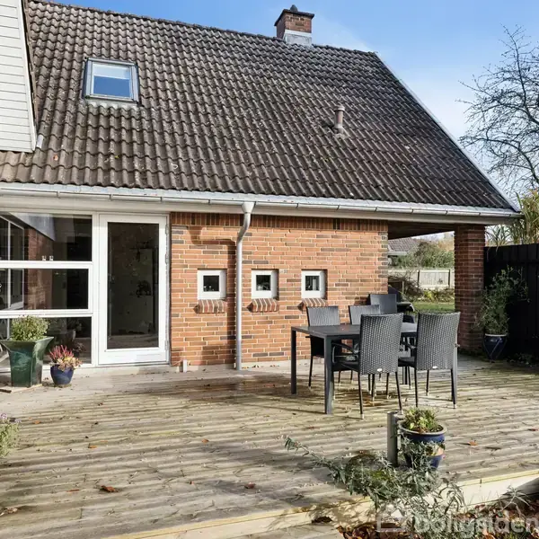 Hus med rødt tegltag og murstensvæg står stille i sollys. En træterrasse med havemøbler og planter skaber en hyggelig udendørs atmosfære. Skyfrit himmel og træer i baggrunden.