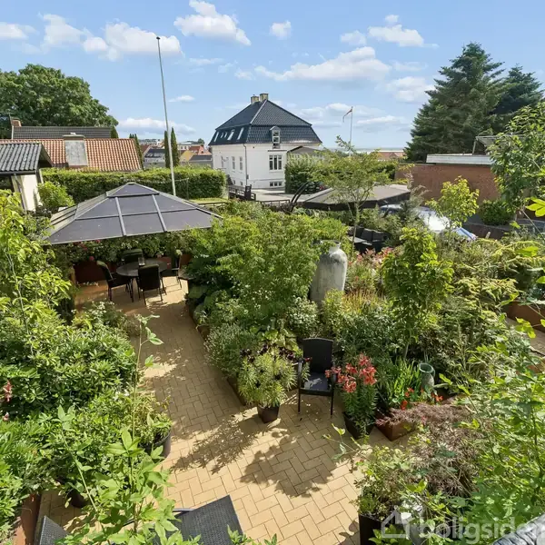 Terrasse med fliser er omgivet af frodige grønne planter og blomster. Et sorthavebord med parasol står under en blå himmel, hvilket skaber en hyggelig, privat udendørs oase.