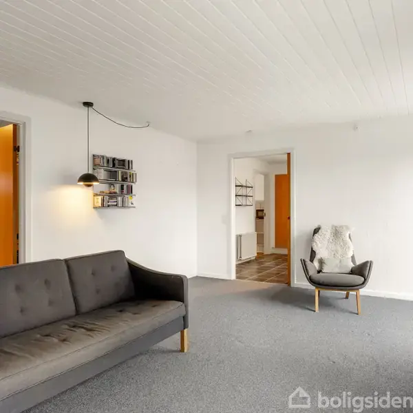 Grå sofa står i et lyst rum med hvide vægge og loft. En moderne lænestol med et lys tæppe står nær en døråbning. Lyskilde og hylder på væggen.