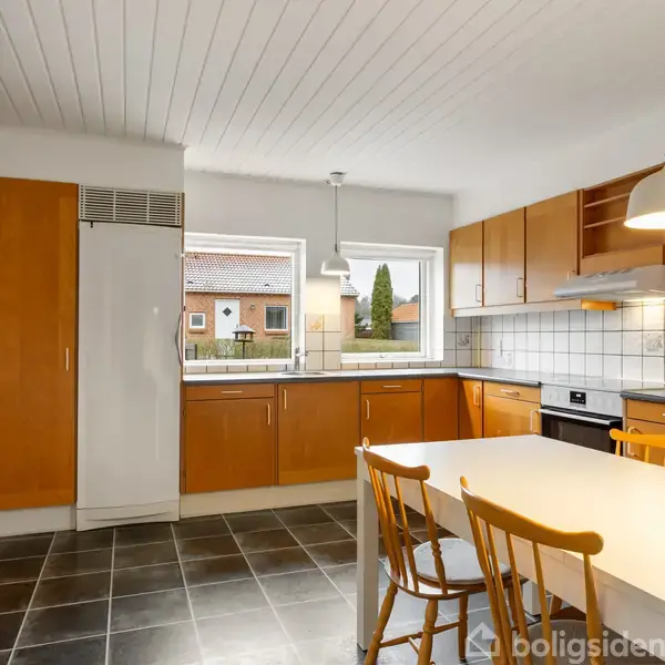 Køkken med lysebrune skabe og hvidt køleskab, stående ved et hvide fliser. Bord med to stole centralt, hængende lampe over. Store vinduer giver udsigt til naboers huse.