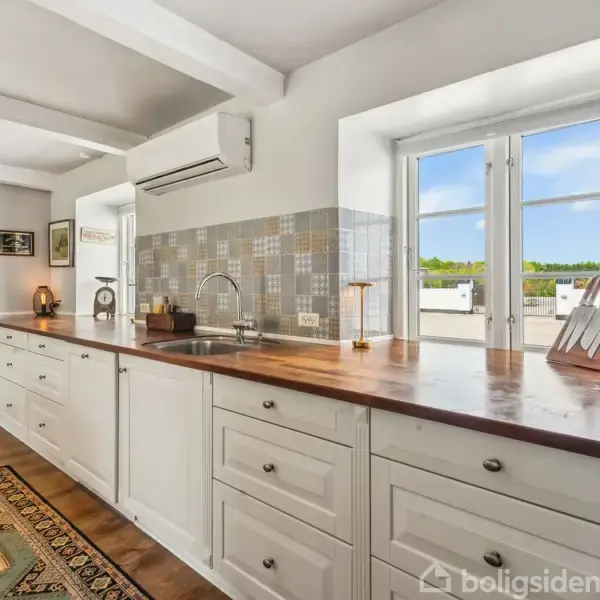 Et køkken med hvide skabe og træbordplader, en vask med en armatur, flere små lamper, et tæppe på gulvet, og vinduer med udsigt til en grøn have.