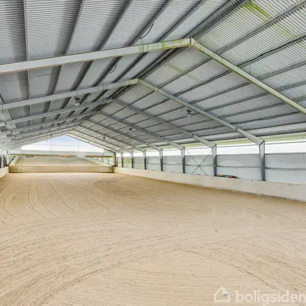 En stor, overdækket ridehal med sandbund, omringet af høje mure. Taget består af metalpaneler, og der er naturligt lysindfald fra vinduer langs siderne.
