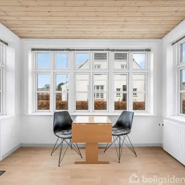 To stole og et bord står i et lyst rum med store vinduer. Udenfor ses hække og nabohuse. Gulvet er i lyst træ, og loftet er træbeklædt.