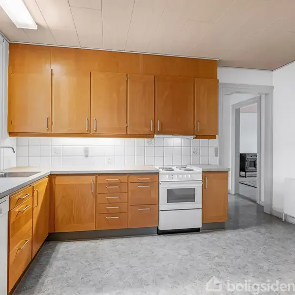 Et køkken med træskabe og hvide fliser. En opvaskemaskine og komfur står langs væggen. Store vinduer giver naturligt lysindfald. En dør fører til et tilstødende rum.