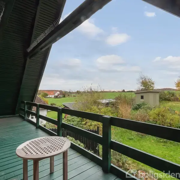 Balkon med trægulv og møbler, som står stille. Udsigt over en grøn have med en grå skur, marker og huse i baggrunden under en blå himmel med skyer.