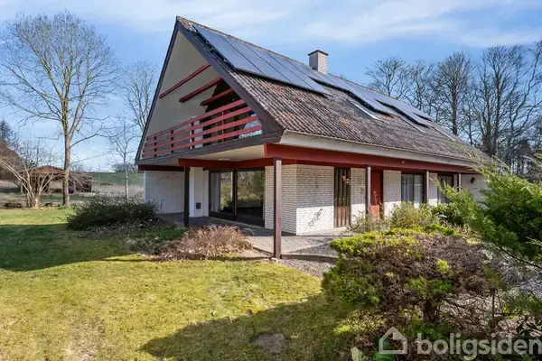 En moderne villa med solpaneler på taget står i en grøn have omgivet af træer. Huset har store vinduer og en balkon med rødt rækværk.