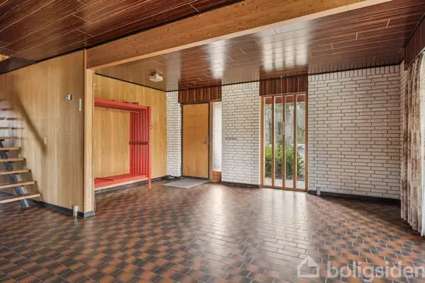 Trægulv med ternet mønster strækker sig gennem en åben entré med hvide murstensvægge og træloft. En rød garderobe og en trætrappe fører til en øverste etage.