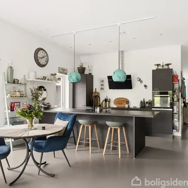 Køkken med en moderne ø med barstole, omgivet af hylder og et spisebord med blå stole. Lysekroner hænger over øen, og en stor ur er på væggen.