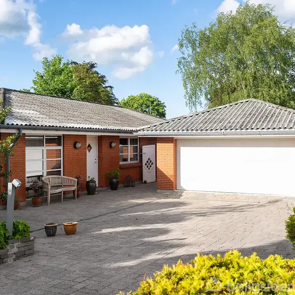 Et rødt murstenshus med en flisebelagt indkørsel, der fører til en hvid garageport. Forhaven har grønne buske og potteplanter, og der står en bænk ved hoveddøren.