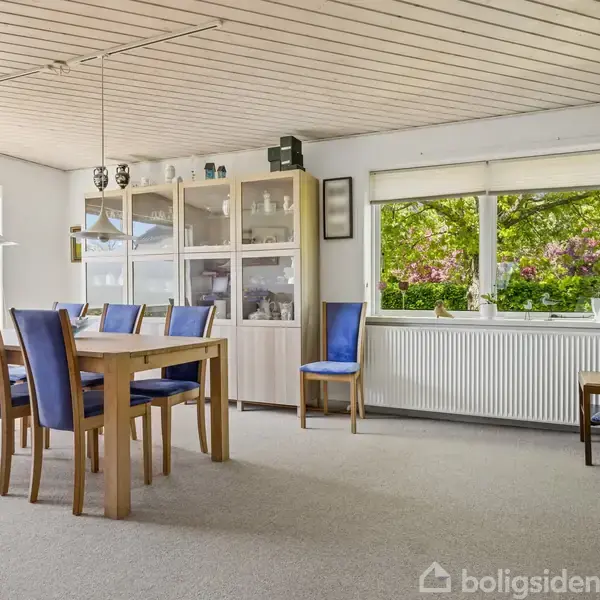 Spisebord med seks blåpolstrede stole står i en lys spisestue. Skab fyldt med porcelæn i baggrunden. Store vinduer giver udsigt til grønne træer og blå himmel udenfor.