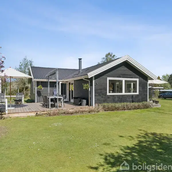 Terrasse med havemøbler og en parasol står på et trædæk, omgivet af en frodig have med buske og træer under en klar blå himmel. En grill står til højre.