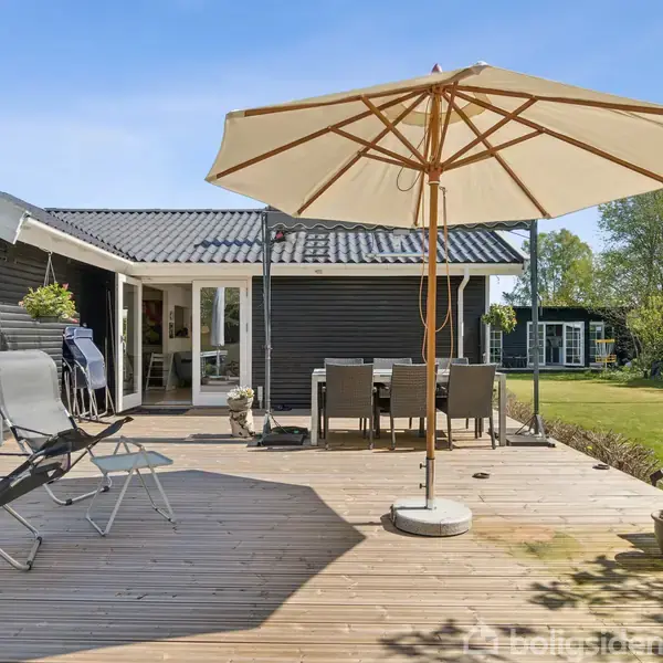 Terrasse med to liggestole og et bord med stole under en parasol. Huset er mørkt med hvide døre, omgivet af en velholdt have og blå himmel.