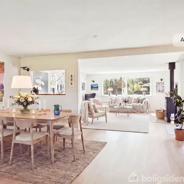 En lys spisestue viser et træbord med stole, prydet med blomster og stearinlys. I baggrunden ses en stue med sofaer ved et stort vindue. Tekst: "AI visualisering".